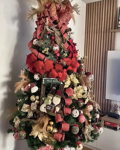 A mesma árvore em 2025, com laços vermelhos, plumas douradas e placa de Feliz Natal