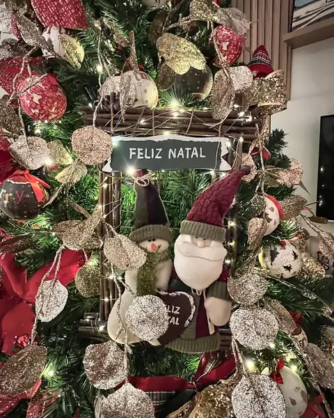 Detalhe da árvore com placa de Feliz Natal, bonecos de pano e folhas prateadas