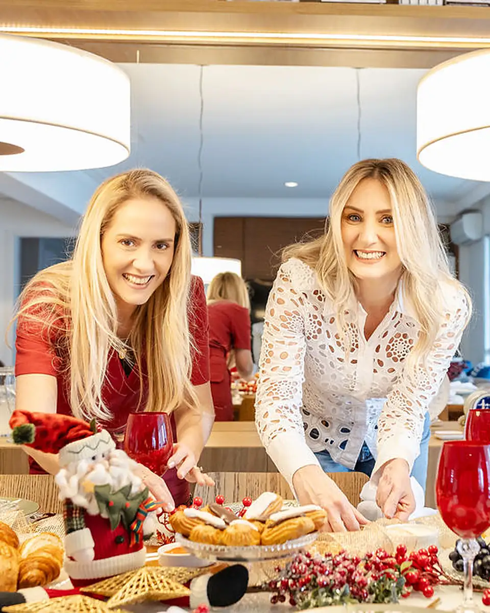 Margot, de vermelho, e Andressa, de blusa de renda branca, sorrindo à mesa posta para o Natal, com taças vermelhas e doces